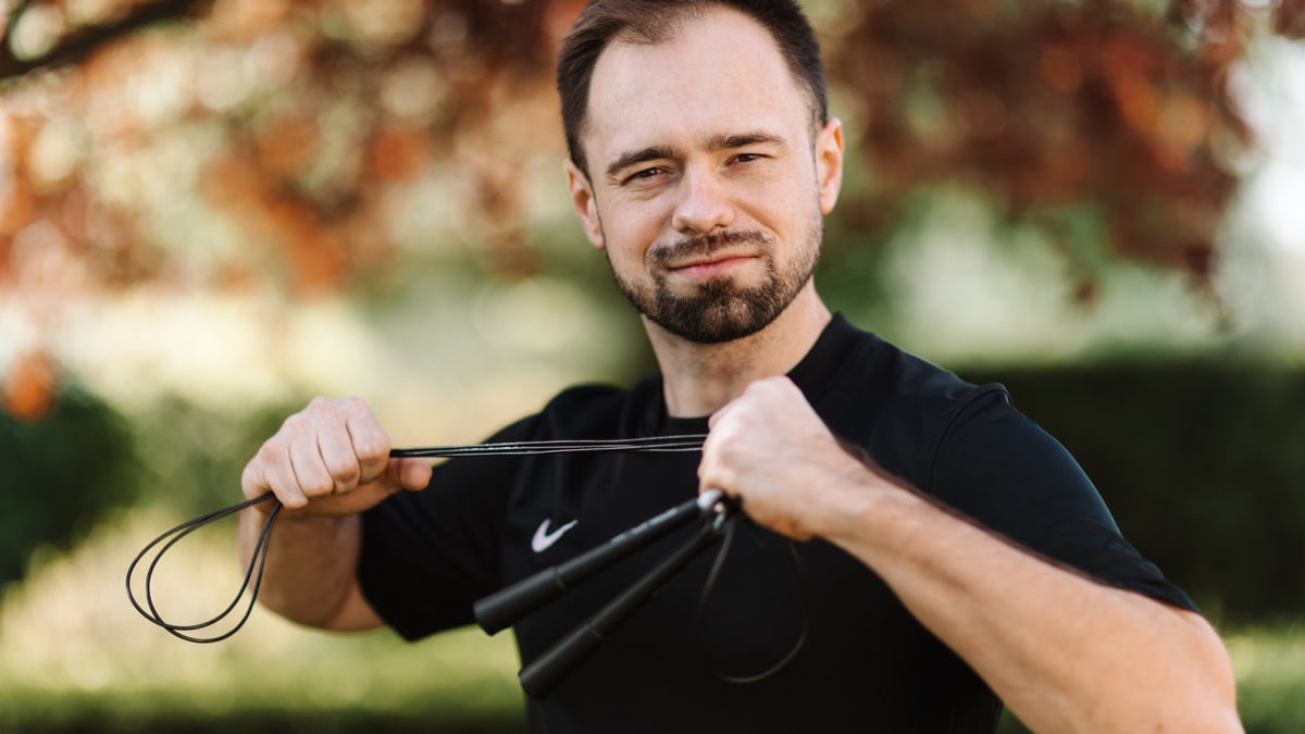 Bearded man in a black shirt confidently posing with a jump rope outdoors.